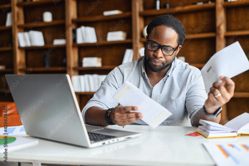 Worried African-American looking through correspondence, a guy holds envelopes in hands, got fine by post, concentrated man sitting at the desk with a laptop in contemporary office Worried African-American looking through correspondence, a guy holds envelopes in hands, got fine by post, concentrated man sitting at the desk with a laptop in contemporary office
