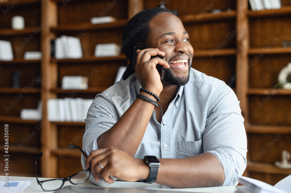 © Vadim Pastuh - Cheerful African-American male employee wearing smart casual shirt holding phone conversation, a man sitting at the desk in modern office, a biracial entrepreneur talking over smartphone and laughs