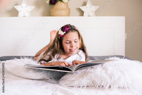Little girl reads a book on the bed in a bright room. The child lies in the bedroom with a book and enthusiastically studies the book. Free space for text. High quality photo