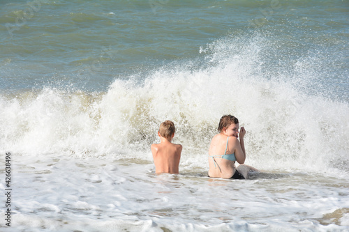 Two children sit laughing on the beach in the waves