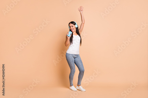 Full size photo of optimistic brunette girl dance with headphones in telephone wear white top jeans sneakers isolated on beige background © deagreez