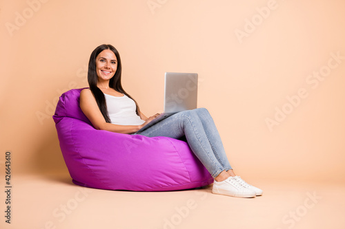 Full size profile photo of optimistic brunette girl sit pouf type laptop wear white top jeans sneakers isolated on beige background photo
