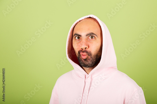 Young bald caucasian man in pink hoodie isolated on green background funny doing grimaces photo