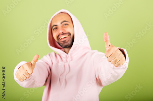 Young bald caucasian man in pink hoodie isolated on green background happy doing gesture success excited shows thumbs up