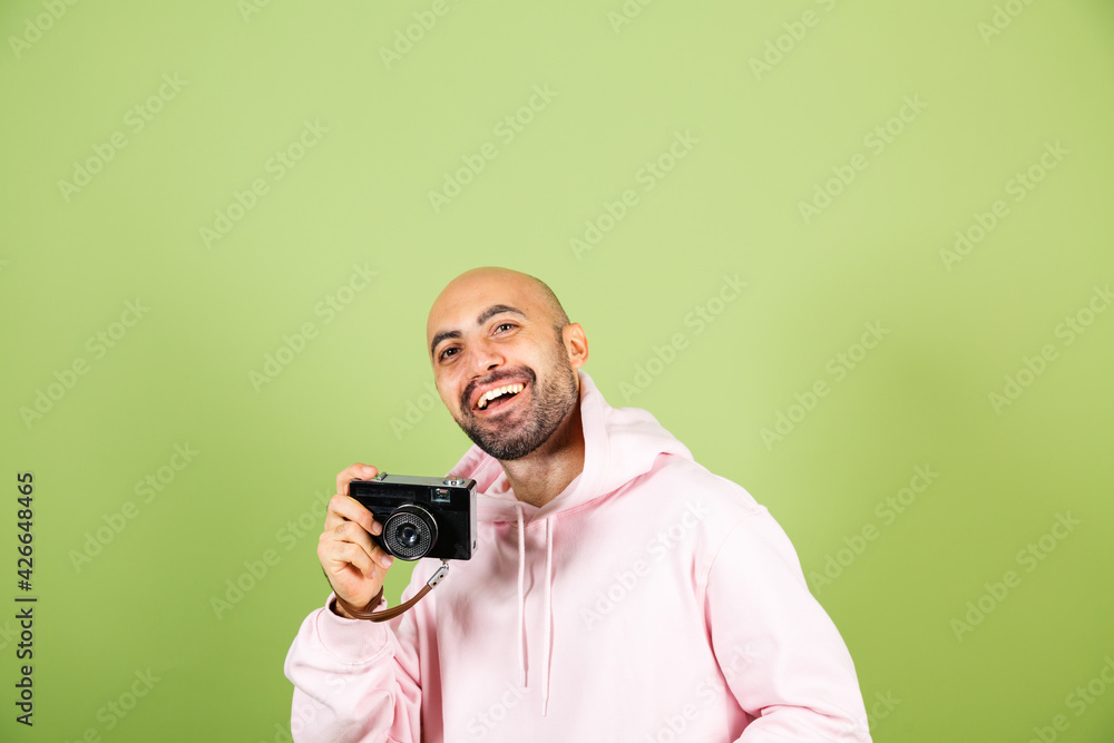 Young bald caucasian man in pink hoodie isolated on green background positive hipster hold camera Young bald caucasian man in pink hoodie isolated on green background positive hipster hold camera
