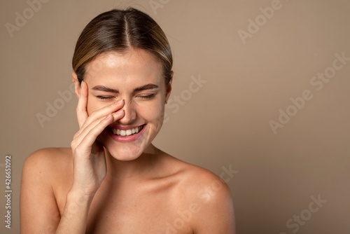Skin care. Woman with beauty face touching healthy facial skin. Beautiful smiling Caucasian female model with natural makeup touching glowing hydrated skin on beige background