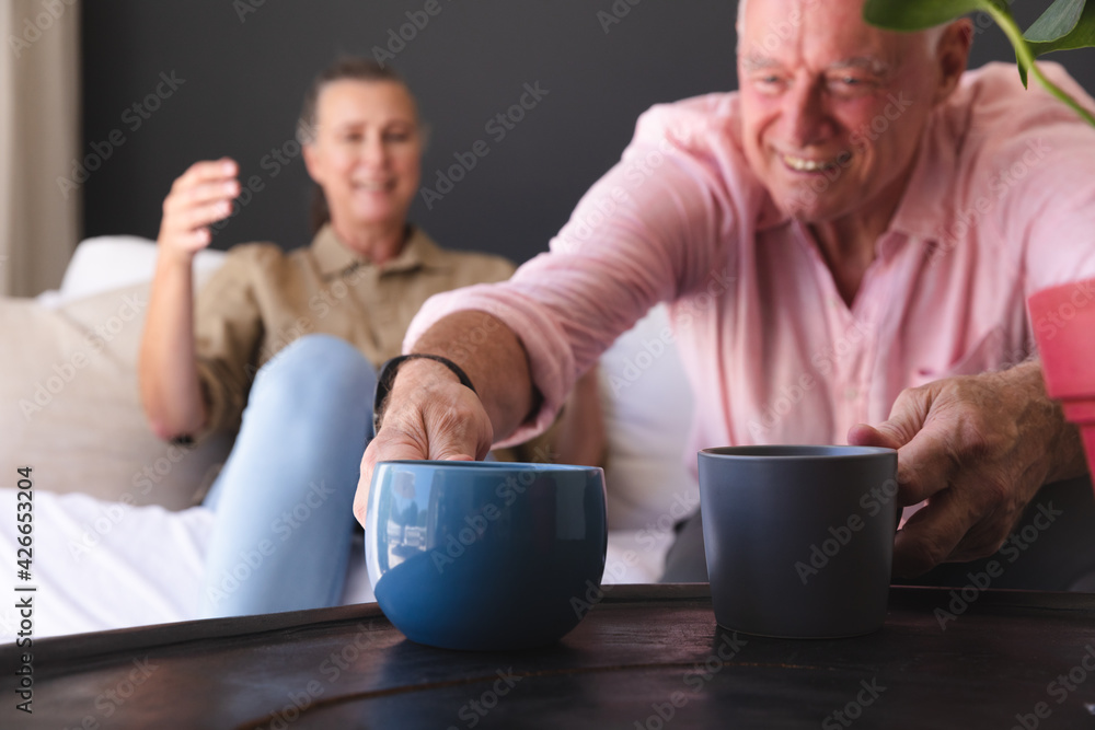 Happy caucasian senior couple relaxing in living room, man putting cups on table and smiling Happy caucasian senior couple relaxing in living room, man putting cups on table and smiling
