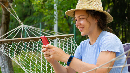 Beautiful young curly woman looking at mobile phone, sitting relaxing in hammock in green garden outdoors. Read news,sharing data on social media,chatting,shopping online,booking, making order in app