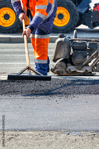 A road worker levels the fresh asphalt with a wooden level on a section of the road against the background of road machinery.