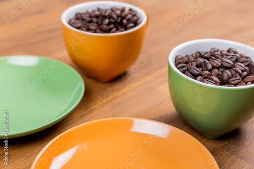 coffee composition, close-up of two cups of coffee one orange and one green with their plates full of coffee beans on a wooden surface, horizontal photo