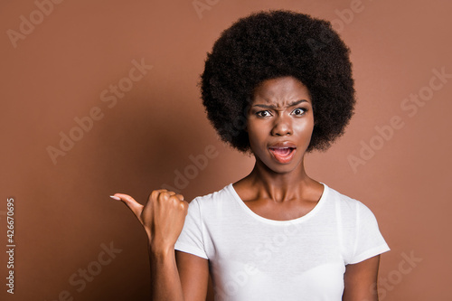 Photo of questioned attractive dark skin girl open mouth indicate thumb empty space isolated on brown color background