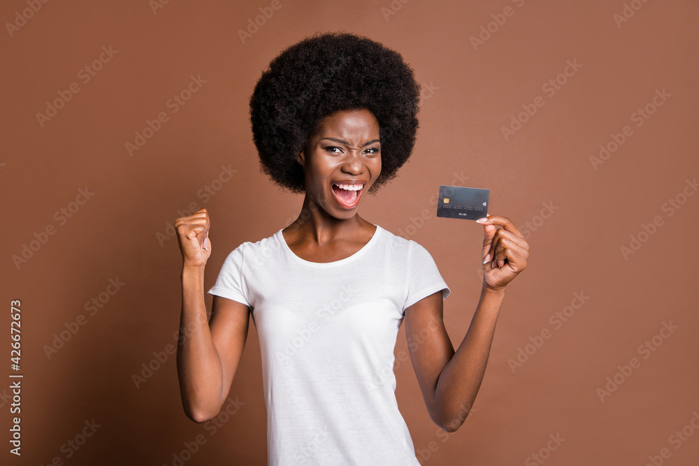 Portrait of astonished dark skin girl hand hold debit card open mouth shout fist up isolated on brown color background Portrait of astonished dark skin girl hand hold debit card open mouth shout fist up isolated on brown color background