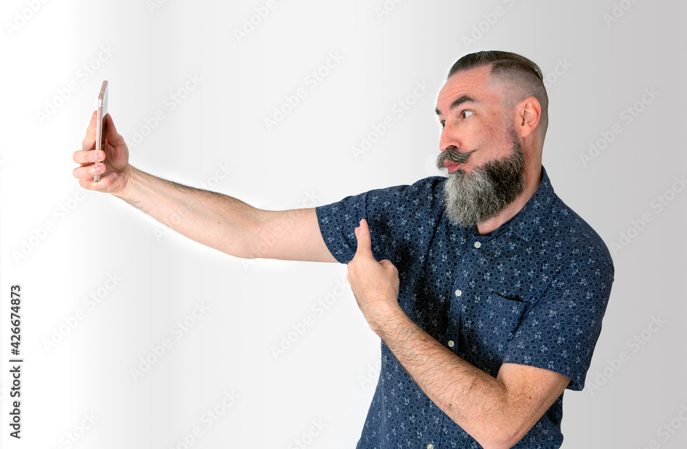 Caucasian Hipster 40-45 years old with big gray half beard putting grimaces and victory sign towards his smart phone to make a selfie or video call on White background Caucasian Hipster 40-45 years old with big gray half beard putting grimaces and victory sign towards his smart phone to make a selfie or video call on White background