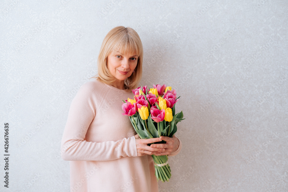 Portrait of Beautiful mature happy smiling woman with blonde hair, holding bouquet of pink and yellow tulip flowers in knitted bag. White background, copy space, close up. Portrait of Beautiful mature happy smiling woman with blonde hair, holding bouquet of pink and yellow tulip flowers in knitted bag. White background, copy space, close up.
