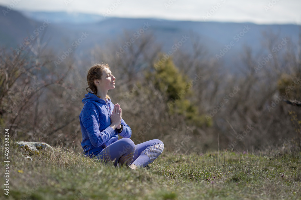 A girl in sports clothes sits on a mountain in the lotus position and meditates, does breathing exercises
