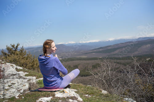 A girl in sports clothes sits on the top of a mountain in a lotus position and admires the beautiful view with her back to the camera and meditates