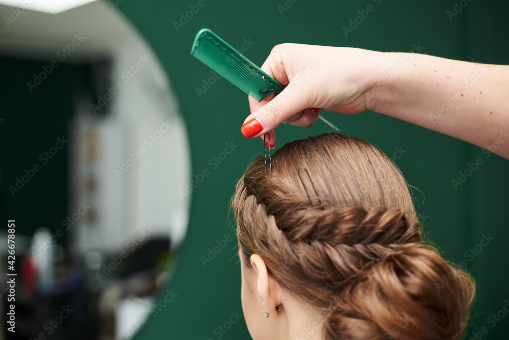 Fototapeta premium Professional hairdresser making coiffure for female client in front of big mirror. Work process in beauty studio. Bride getting ready for wedding in beauty salon. Close-up picture of fancy hairdo.