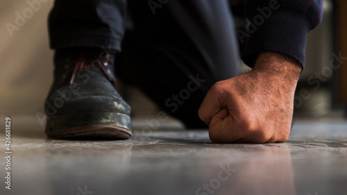 Close up detail of a crouched unrecognizable man's fist and foot with boot, standing on gray, screed floor in front shot.