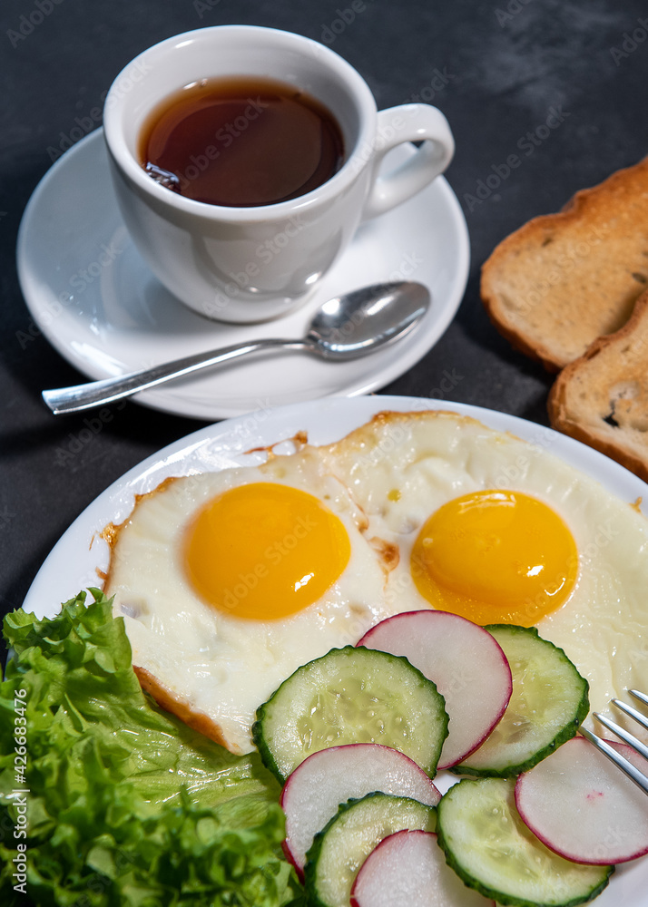 Two fried eggs, lettuce, cucumber and radish slices on a white plate, toast and a cup of tea or coffee. Scrambled eggs. Breakfast. Healthy eating. Close-up Two fried eggs, lettuce, cucumber and radish slices on a white plate, toast and a cup of tea or coffee. Scrambled eggs. Breakfast. Healthy eating. Close-up