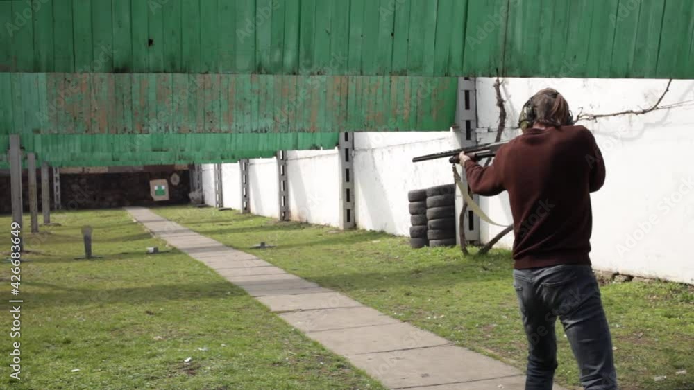 A young man shoots a pump-action firearm at flags, targets. A man in protective glasses and headphones in a dash. A green field with bullet protection outdoor. Sports shooting, weapons hobby. shotgun A young man shoots a pump-action firearm at flags, targets. A man in protective glasses and headphones in a dash. A green field with bullet protection outdoor. Sports shooting, weapons hobby. shotgun