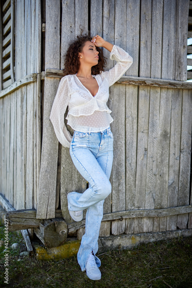 A beautiful woman with curly hair poses near an old wooden house. fashion style.