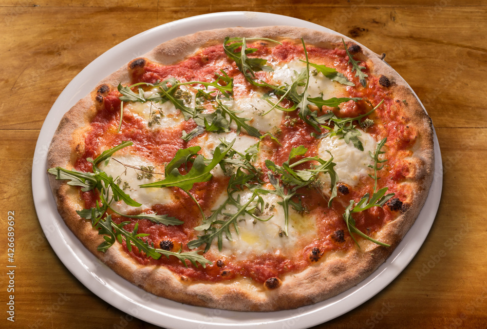 Pizza with arugula leaves, tomato sauce, mozzarella cheese in white plate on wooden table, top view Pizza with arugula leaves, tomato sauce, mozzarella cheese in white plate on wooden table, top view