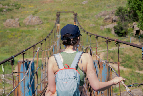 A brunette girl, a Caucasian in a cap, is walking on a suspension bridge. Backpack at the back. Rocky shore in the distance