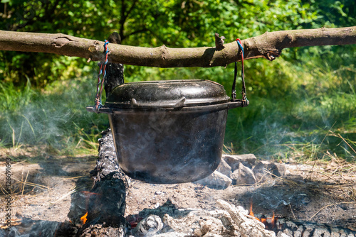 A pot for cooking outdoors is hanging on a stick. Logs smolder underneath and smoke is visible. The background is blurred. Field kitchen