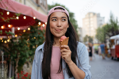 Portrait of beautiful asian woman eating ice cream on the street.  Emotional hipster wearing casual clothing holding tasty summer dessert looking away outdoors. Food festival