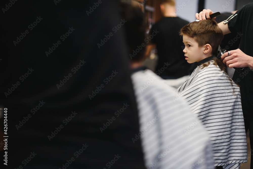 Fototapeta premium A little cute boy sits in a hairdresser's at the stylist's, a schoolchild is getting hair cut in a beauty salon, a child at a barbershop's, a short men's haircut.