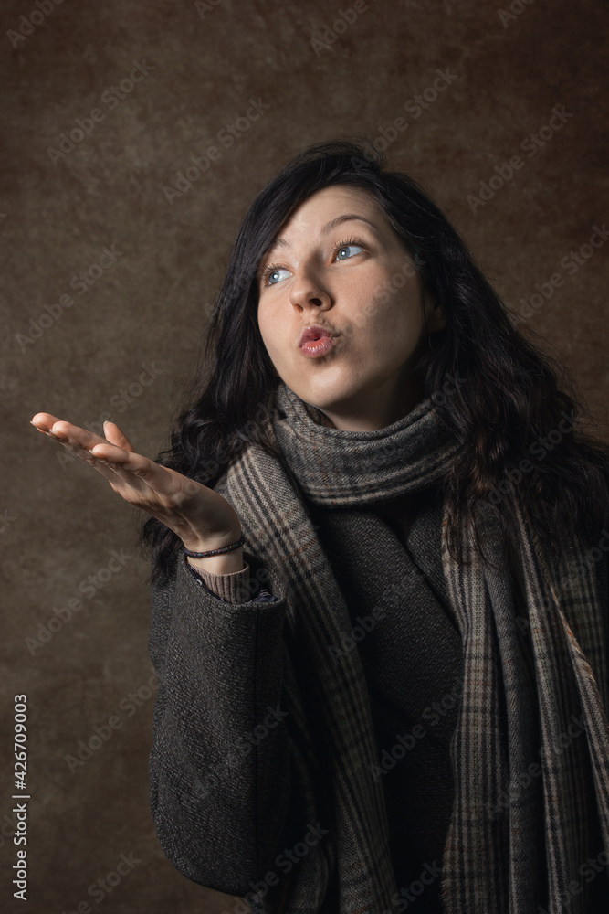 The girl sends an air kiss. Portrait of a girl with a scarf and a coat. Woman portrait on brown background The girl sends an air kiss. Portrait of a girl with a scarf and a coat. Woman portrait on brown background