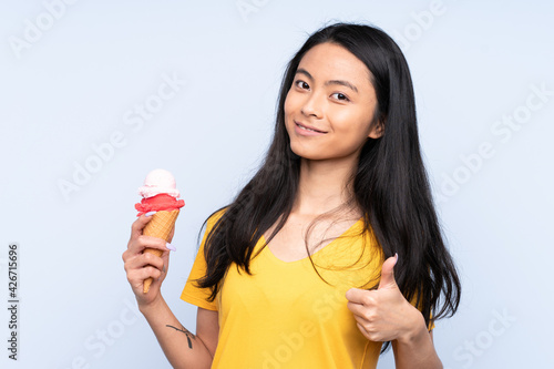 Teenager Asian girl with a cornet ice cream isolated on blue background with thumbs up because something good has happened © luismolinero