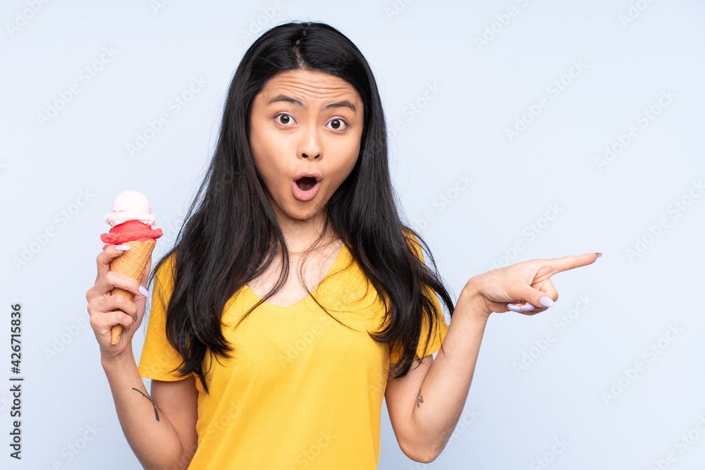 Teenager Asian girl with a cornet ice cream isolated on blue background surprised and pointing side Teenager Asian girl with a cornet ice cream isolated on blue background surprised and pointing side