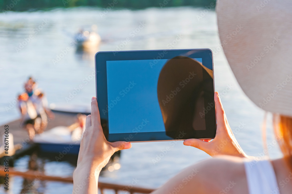 Mock-up of a tablet, a view over the shoulder in the hands of a girl in a hat. Against the background of a wooden embankment with water and a boat.