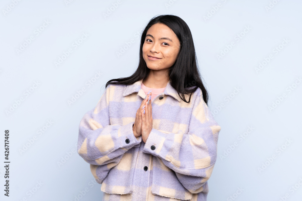 Teenager Chinese woman isolated on blue background keeps palm together. Person asks for something Teenager Chinese woman isolated on blue background keeps palm together. Person asks for something