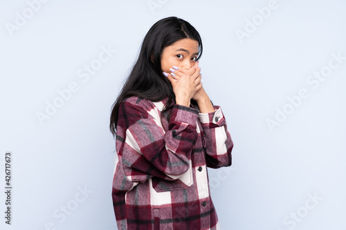 Teenager Chinese woman isolated on blue background covering mouth and looking to the side © luismolinero
