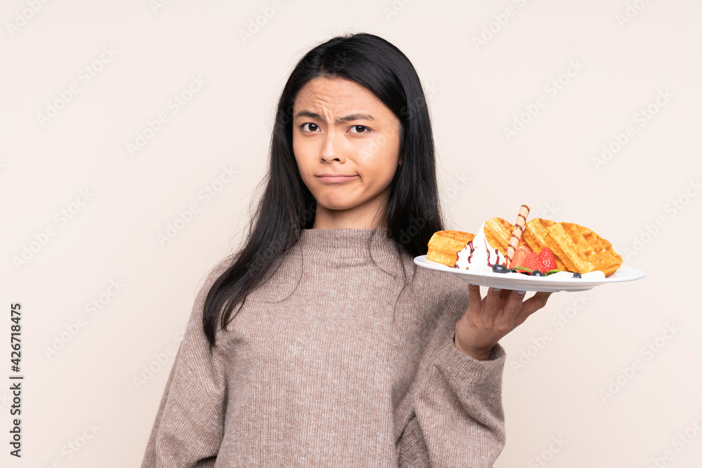 Teenager Asian girl holding waffles isolated on beige background with sad expression Teenager Asian girl holding waffles isolated on beige background with sad expression
