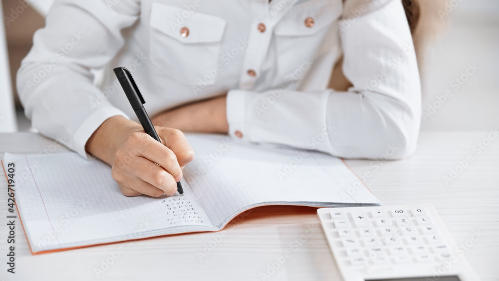 Fototapeta premium Cropped view of girl writing on notebook near calculator on blurred foreground.