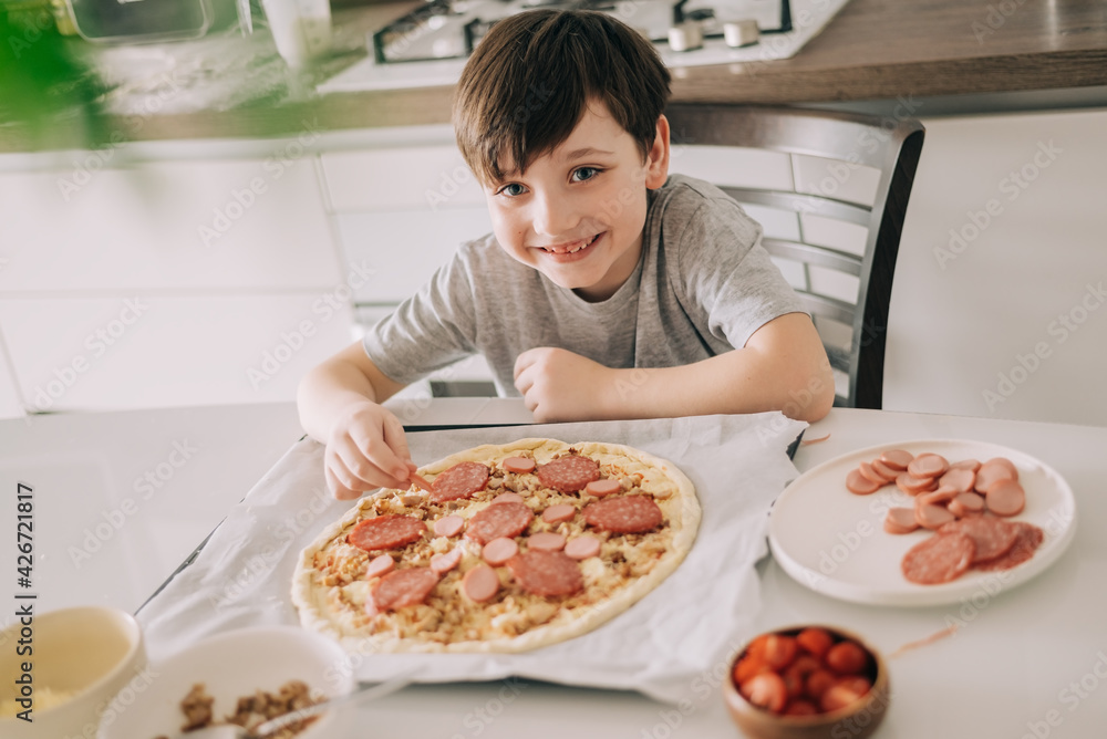 Little kid boy making pizza sitting at the table on the kitchen. Children helping in cooking lifestyle image