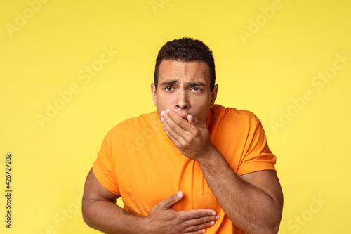 Masculine man want vomit, feeling sick and unwell, ate too much junk unhealthy food, touching stomach and cover mouth with palm as standing yellow background sick, puke from disgust photo