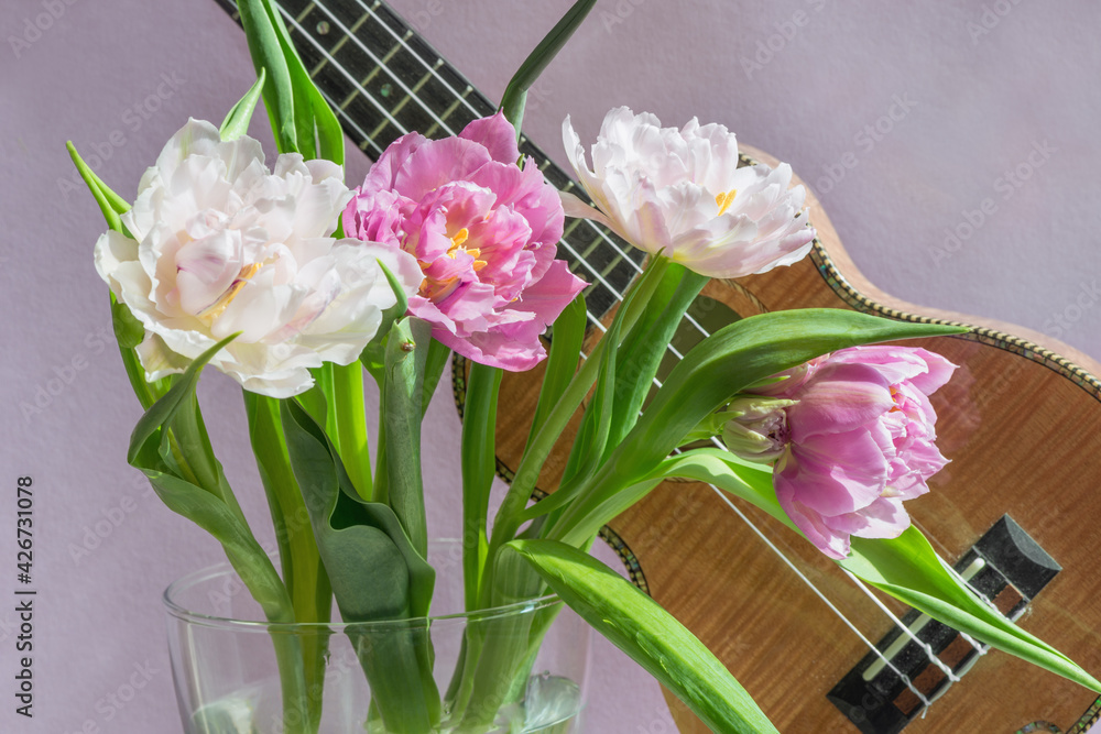 Obraz premium A bouquet of beautiful tulips on a lilac background and a fragment of a musical instrument ukulele.