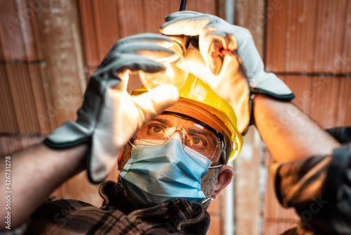 Electrician worker at work replaces the light bulb protected by helmet, safety goggles and gloves. Wear the surgical mask to prevent the spread of Coronavirus. Construction industry. Covid-19 Pandemic