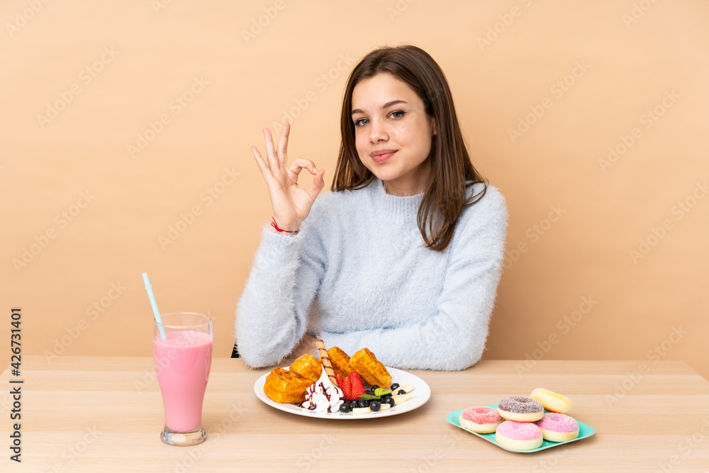 Teenager girl eating waffles isolated on beige background showing ok sign with fingers Teenager girl eating waffles isolated on beige background showing ok sign with fingers