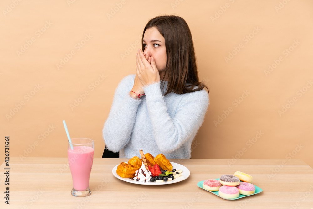 Teenager girl eating waffles isolated on beige background covering mouth and looking to the side Teenager girl eating waffles isolated on beige background covering mouth and looking to the side