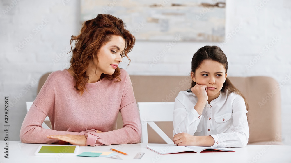 Pensive kid holding pen near notebook and mother during home education. Pensive kid holding pen near notebook and mother during home education.