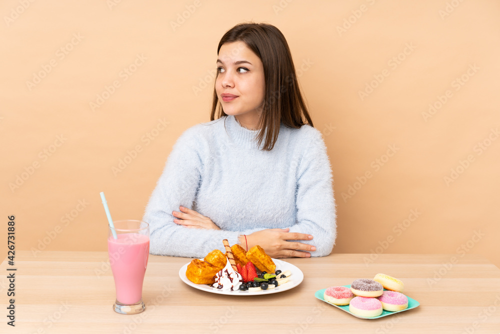 Teenager girl eating waffles isolated on beige background making doubts gesture looking side Teenager girl eating waffles isolated on beige background making doubts gesture looking side