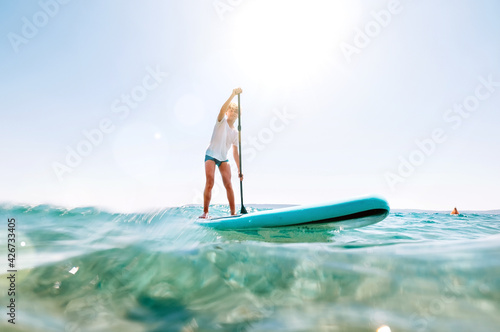 Under the water view angle to the smiling blonde teenager boy rowing stand up paddle board. Active family summer vacation time near the sea concept image.