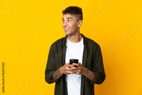 Young brazilian man isolated on yellow background using mobile phone and looking up