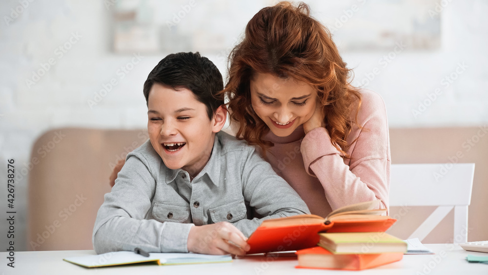 Smiling woman sitting near cheerful son holding book on blurred foreground. Smiling woman sitting near cheerful son holding book on blurred foreground.