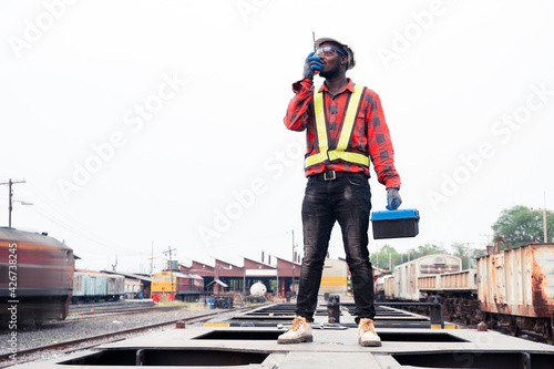 African machine engineer technician wearing a helmet, groves and safety vest is using a wrench to repair the train with talking by radio communication or walkie talkie